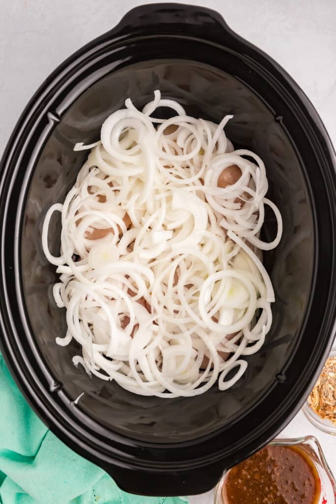 Sliced onions covering raw chicken in a black slow cooker.