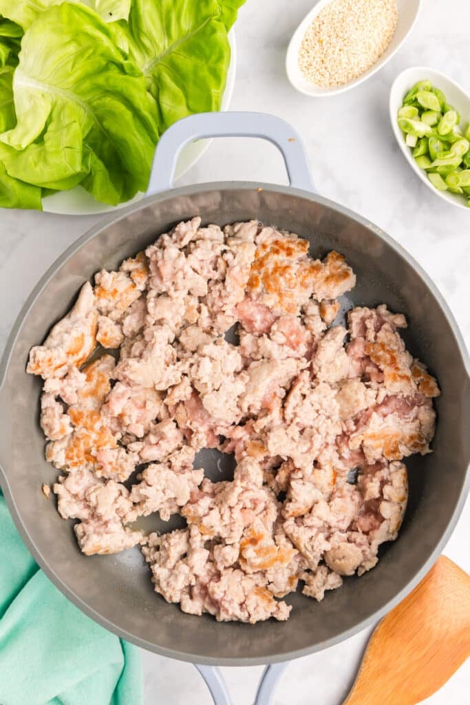 Sautéed ground chicken in a skillet with lettuce leaves nearby.