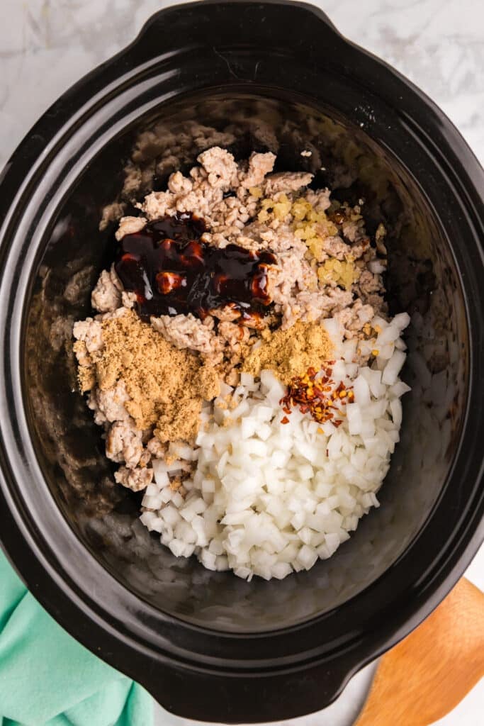 Ground chicken in a slow cooker with diced onions, minced garlic, hoisin sauce, brown sugar, and red pepper flakes before cooking.