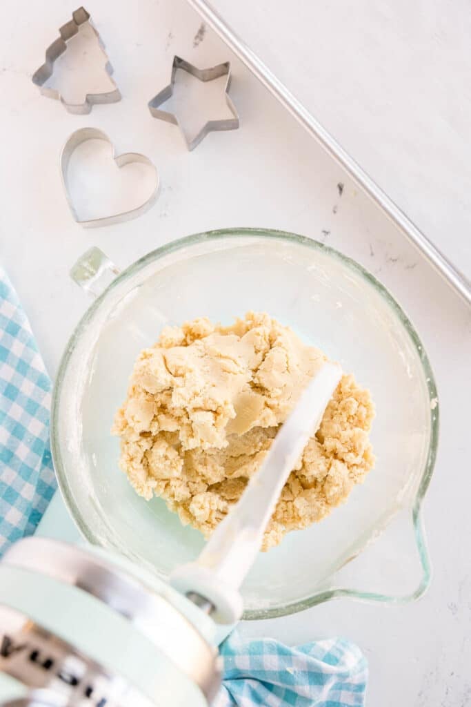 A glass mixing bowl with sugar cookie dough and a stand mixer.