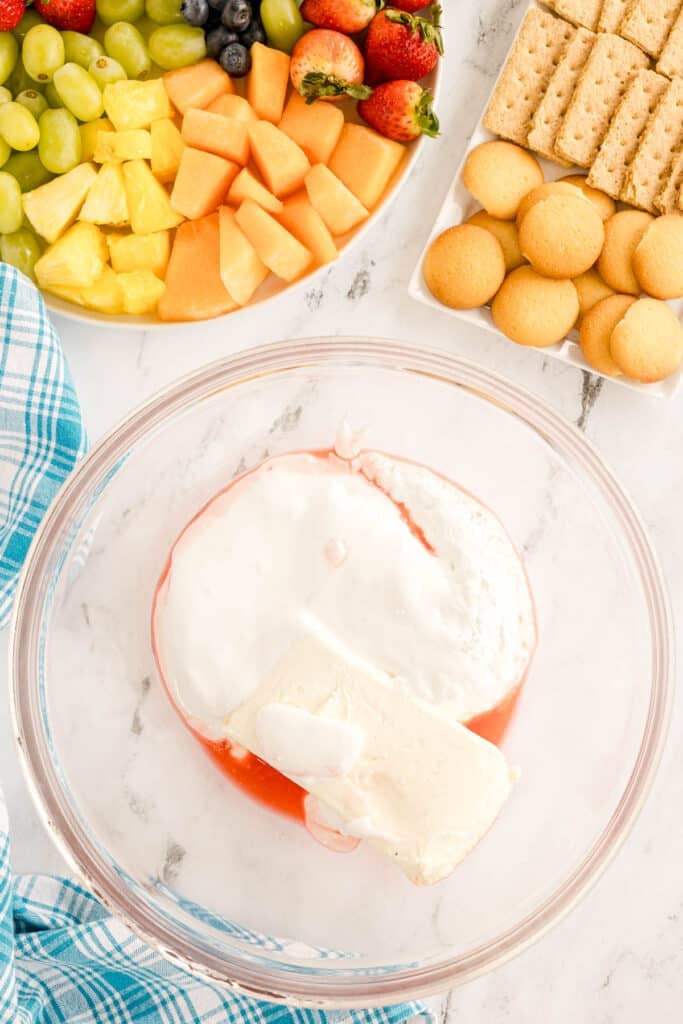 A glass bowl with unmixed fruit dip ingredients, including cream cheese and maraschino cherry juice.