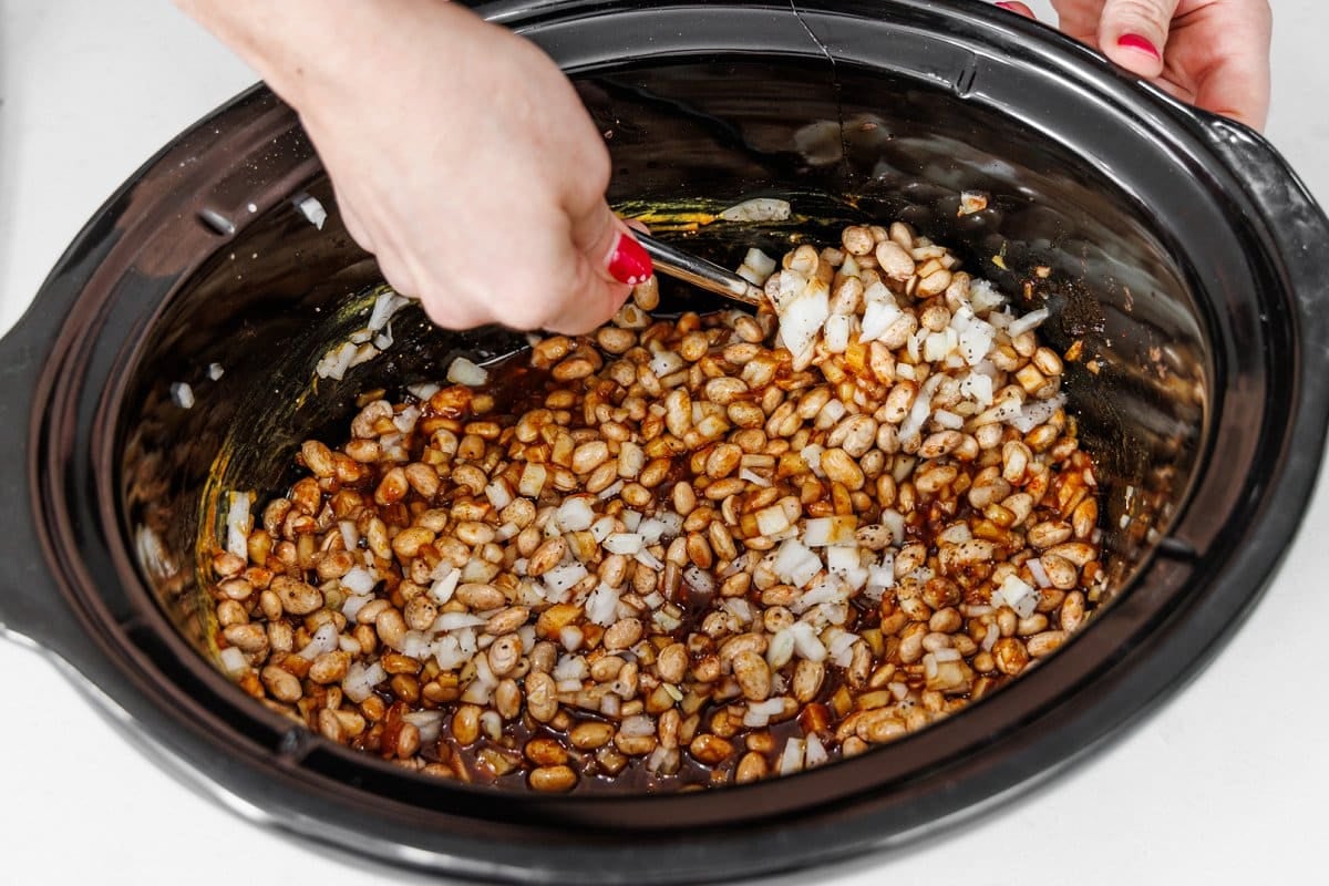 Stirring onions and beans with seasoning in slow cooker bowl.