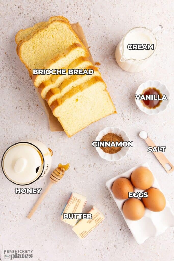 A top-down view of ingredients for French toast, including sliced brioche bread, cream, vanilla, cinnamon, salt, eggs, butter, and honey.