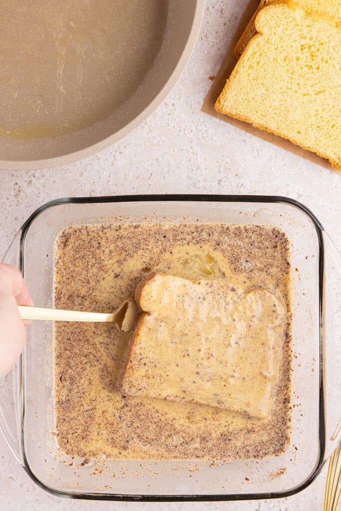 A hand dipping a slice of brioche bread into a cinnamon custard mixture in a glass dish.