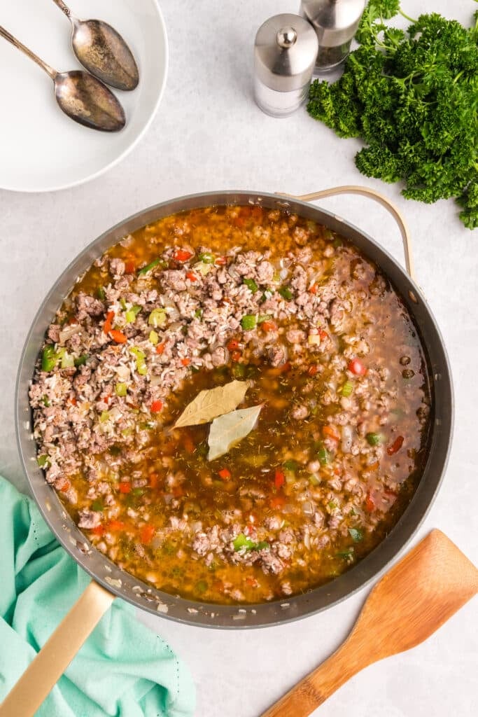 Skillet of dirty rice ingredients simmering with broth and bay leaves.