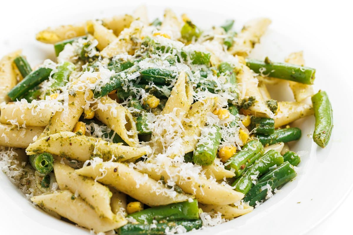 Pesto pasta salad with green beans and corn topped with cheese on a white plate.