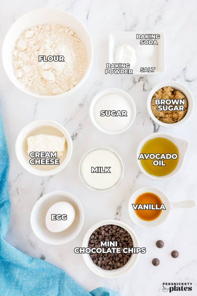 Various labeled baking ingredients in small bowls on a white surface, including flour, sugars, milk, and chocolate chips.