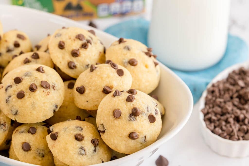 Bowl full of golden mini chocolate chip muffins with a glass of milk and chocolate chips in the background.