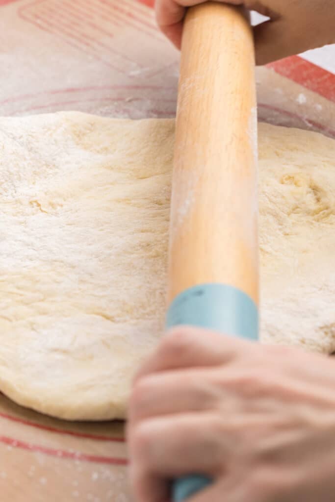 Hands using a rolling pin to flatten dough on a floured surface.