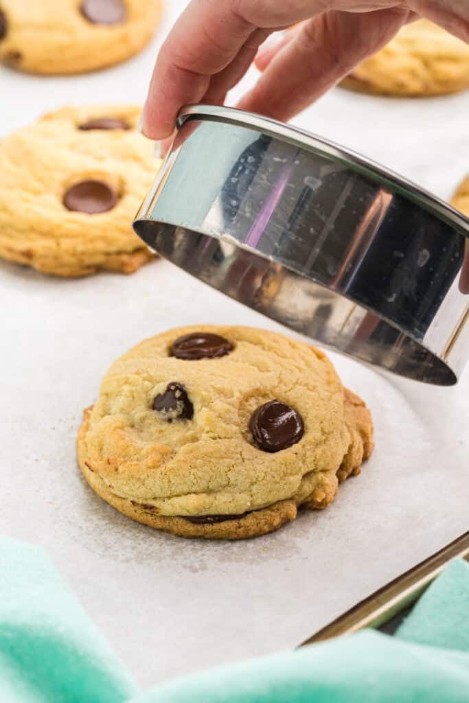 a hand using a circle biscuit cutter to smooth the edges of a chocolate chip cookie.