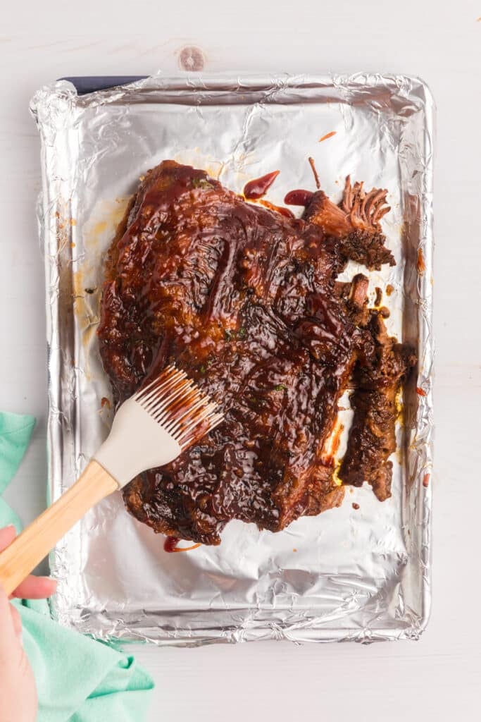 A hand brushes barbecue sauce onto a cooked beef brisket on a foil-lined baking sheet.