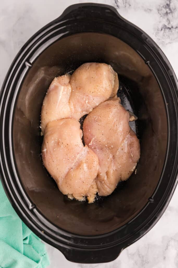 Raw skinless chicken breasts seasoned with salt and pepper are arranged in the bottom of a slow cooker.