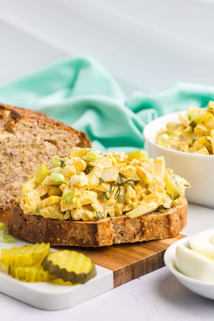 A slice of hearty whole grain bread is topped with a generous serving of creamy dill pickle egg salad, garnished with chopped green onions and fresh dill.