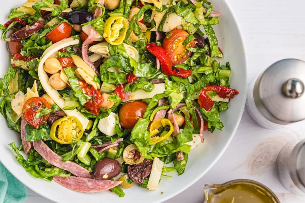 Antipasto salad in a white bowl with pepper mills and dressing.
