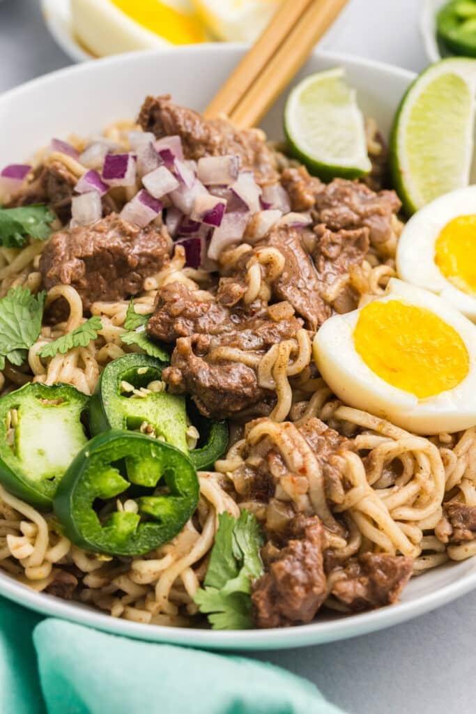 Close-up of birria ramen in a bowl with beef, soft eggs, and jalapeños. Chopsticks rest on the rim of the bowl
