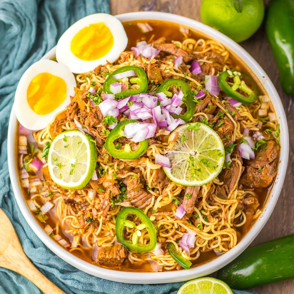 Close-up of birria ramen in a bowl with lime slices, soft eggs, and jalapeños. 