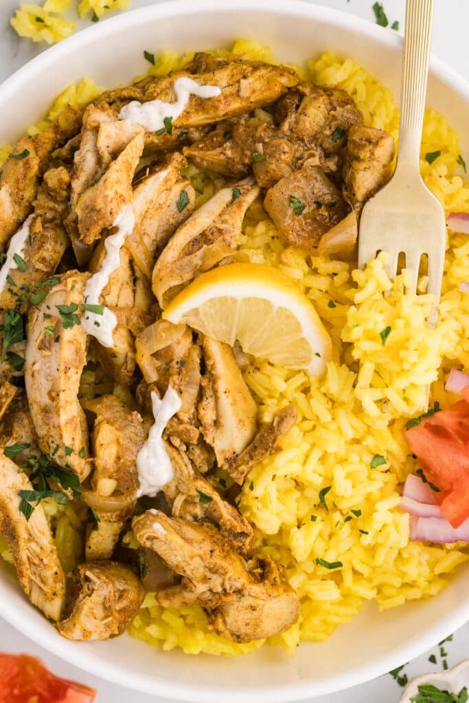 Bowl of yellow rice topped with chicken shawarma, lemon wedge, and vegetables.