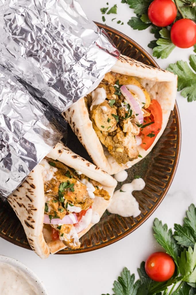 Two chicken shawarma wraps in foil on a plate with tomatoes and parsley.