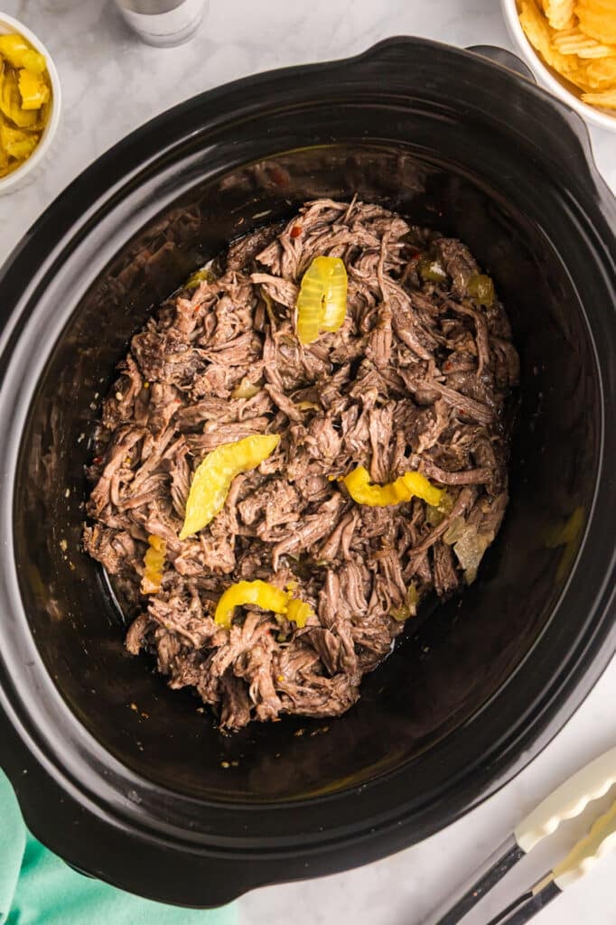Shredded Italian beef with yellow peppers in a black slow cooker.