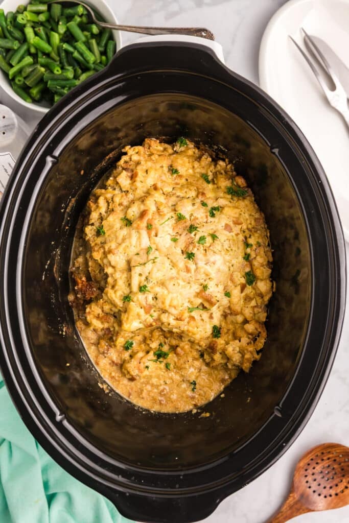 Turkey stuffing casserole in a slow cooker and a bowl of green beans on a white surface.