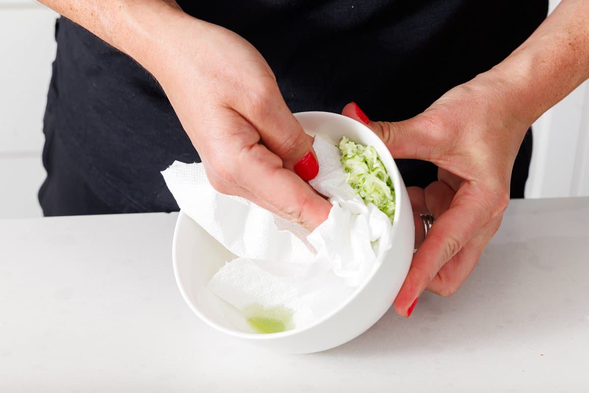 Liz usiing a paper towl to soak up extra moisture from grated cucumber in a bowl.