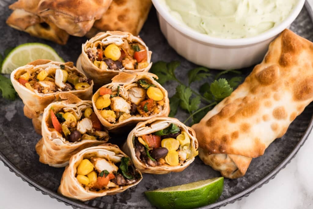 Southwestern egg rolls arranged on a platter with green dip and lime wedges.