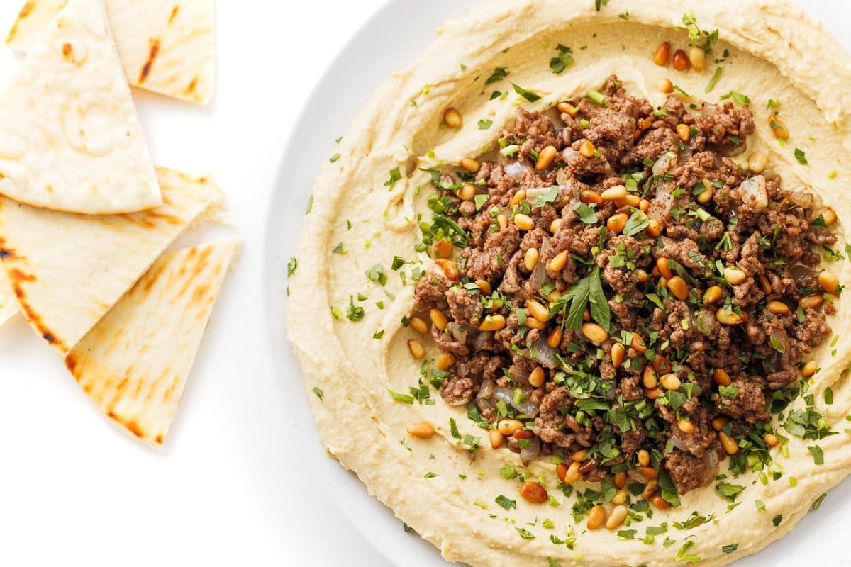Hummus with ground beef and pine nuts on a plate next to pita bread.