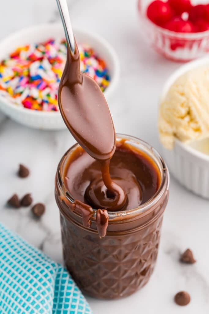 A spoon lifting rich hot fudge from a glass jar.