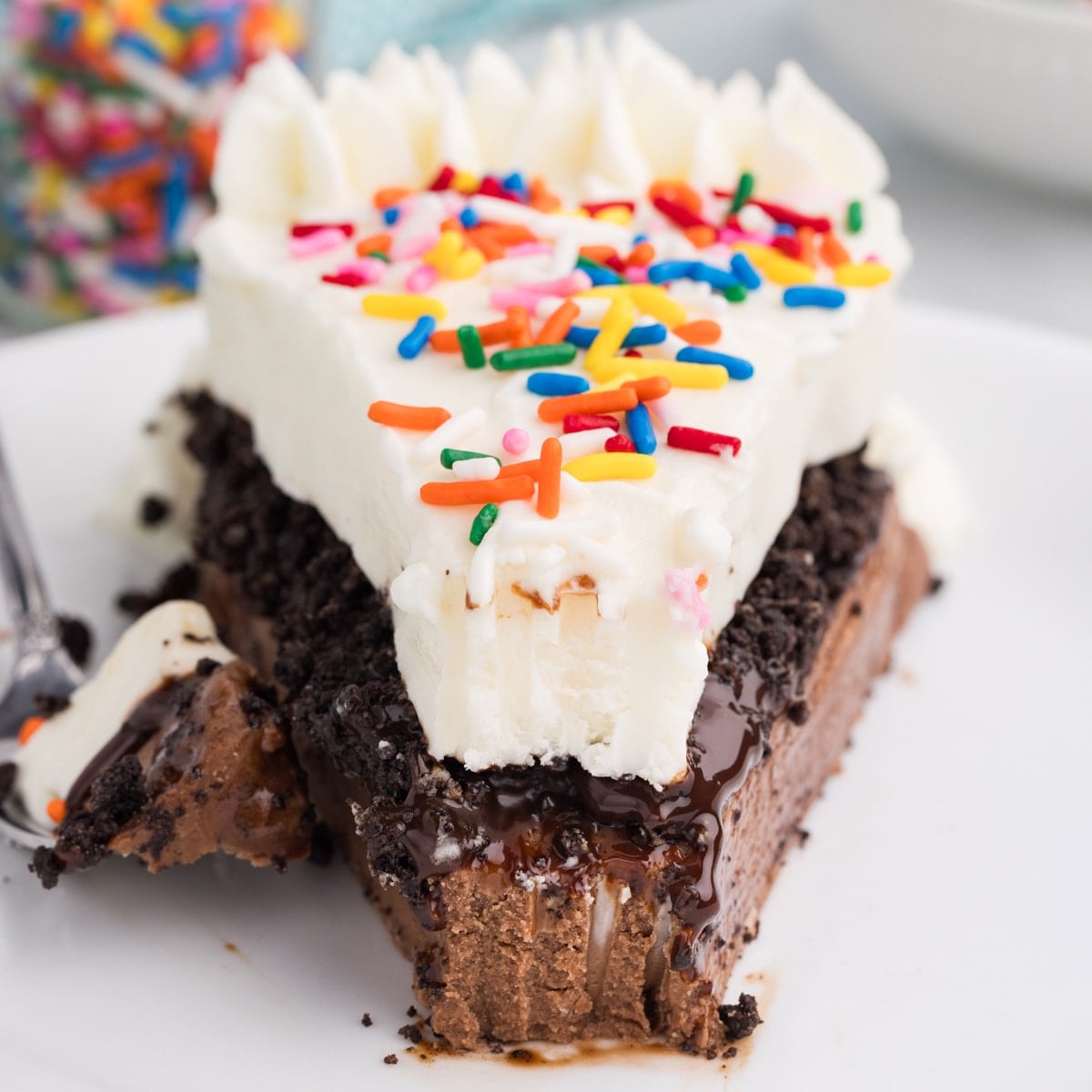 Slice of ice cream cake with white frosting, sprinkles, fudge, and cookie crumble on a plate.