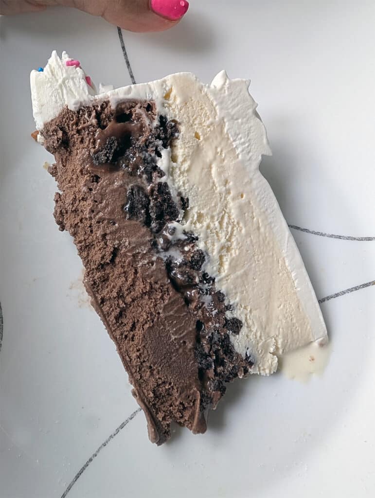 Slice of layered ice cream cake with chocolate and vanilla ice cream, cookie crumble, and fudge on a white plate.
