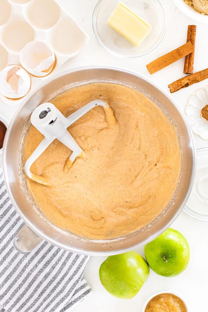 A mixing bowl filled with spiced apple cider donut bread batter on a countertop surrounded by eggshells, a stick of butter, cinnamon sticks, and green apples.