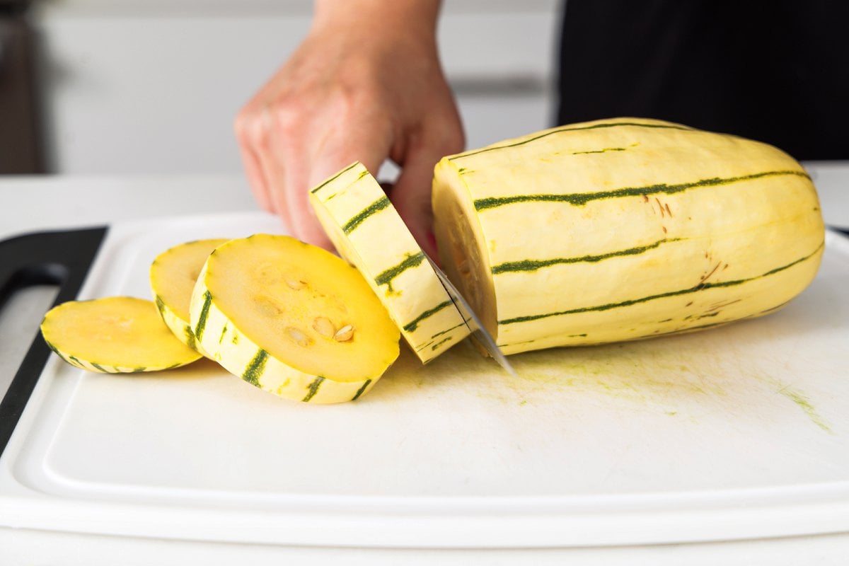 Cutting a delicata squash into slices with a sharp knife on a cutting board.