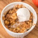 Apple crisp topped with vanilla ice cream in a white ramekin on a wooden board.