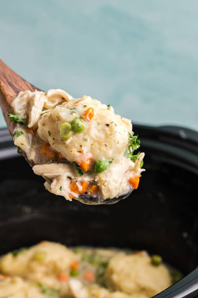 Wooden spoon lifting a dumpling with peas, carrots, and chicken from a slow cooker.