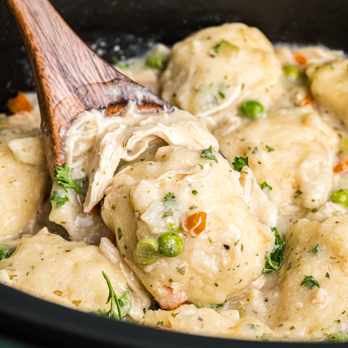 A wooden spoon lifting creamy chicken and dumplings with peas and carrots.