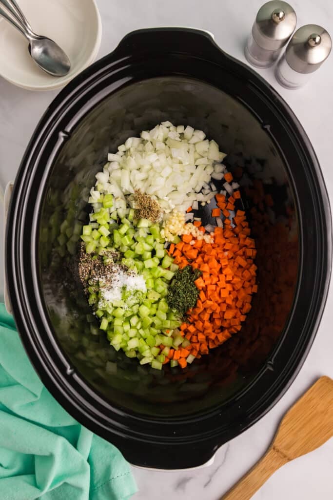Chopped onions, celery, and carrots with herbs and seasoning inside a slow cooker.