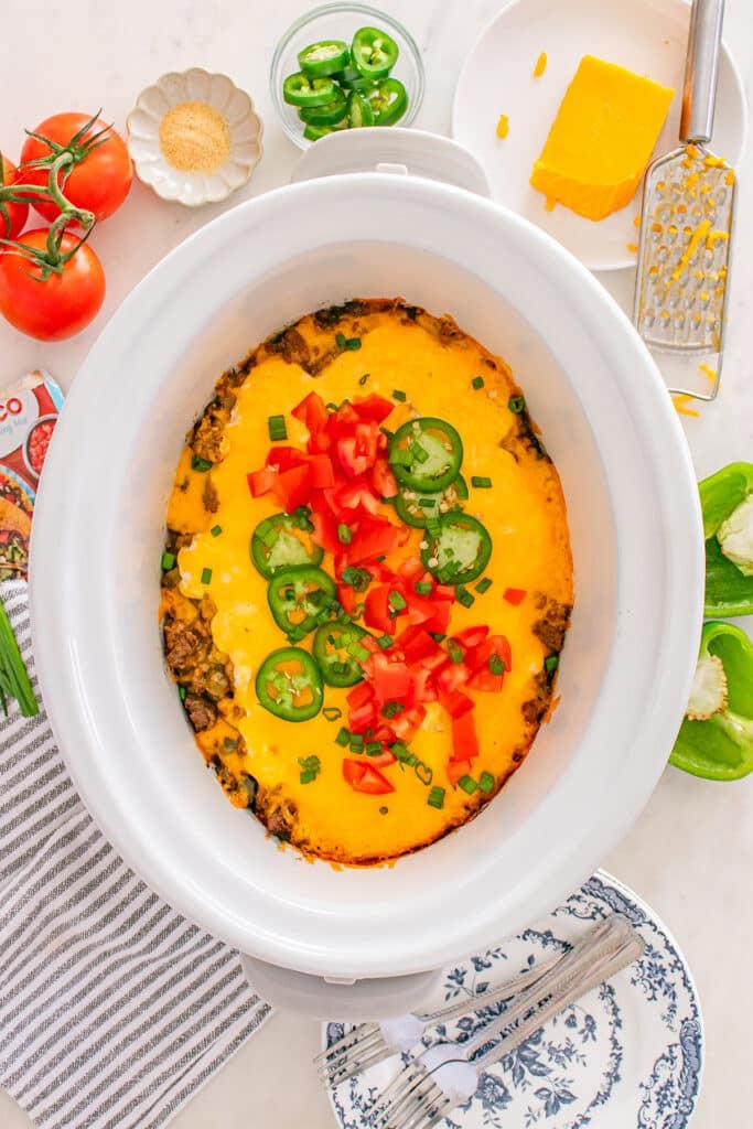 A top-down view of cheesy casserole in a slow cooker topped with tomatoes and jalapeños.