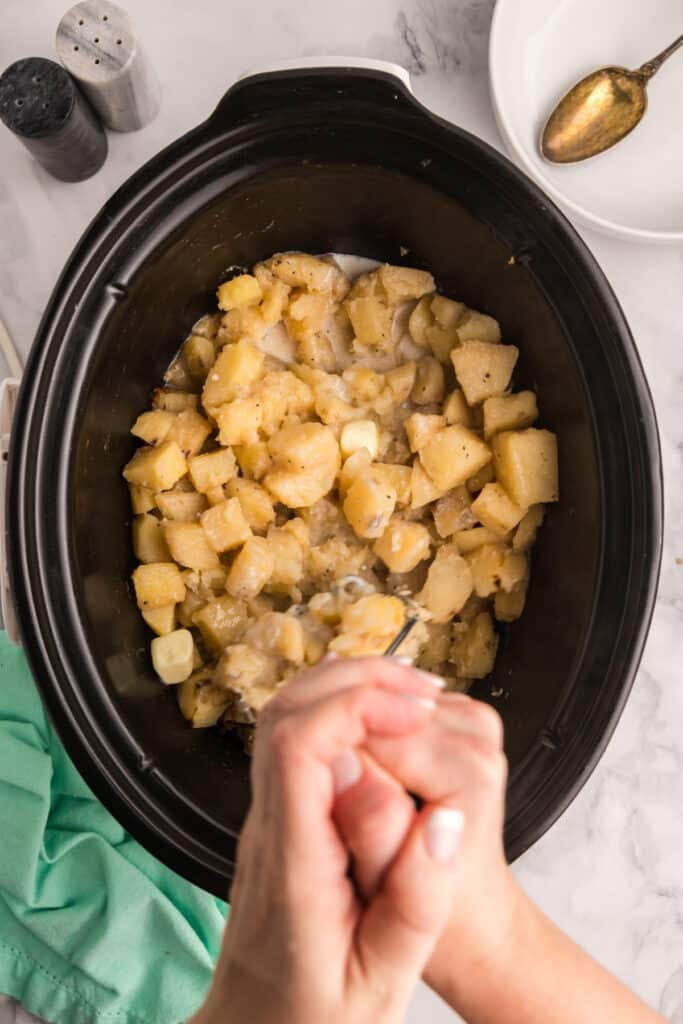 Hands mashing cooked potatoes in a slow cooker.