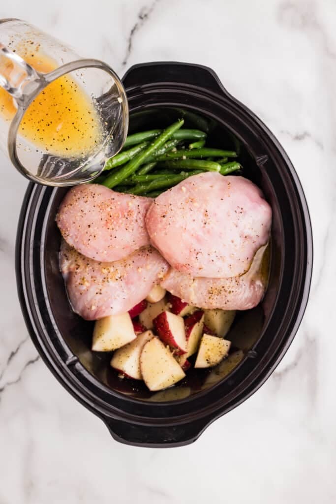 Seasoned liquid being poured over chicken, potatoes, and green beans in a slow cooker.