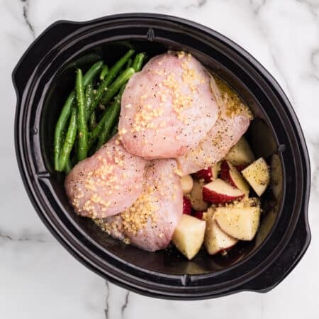 Raw chicken, red potatoes, and green beans in a slow cooker topped with minced garlic.