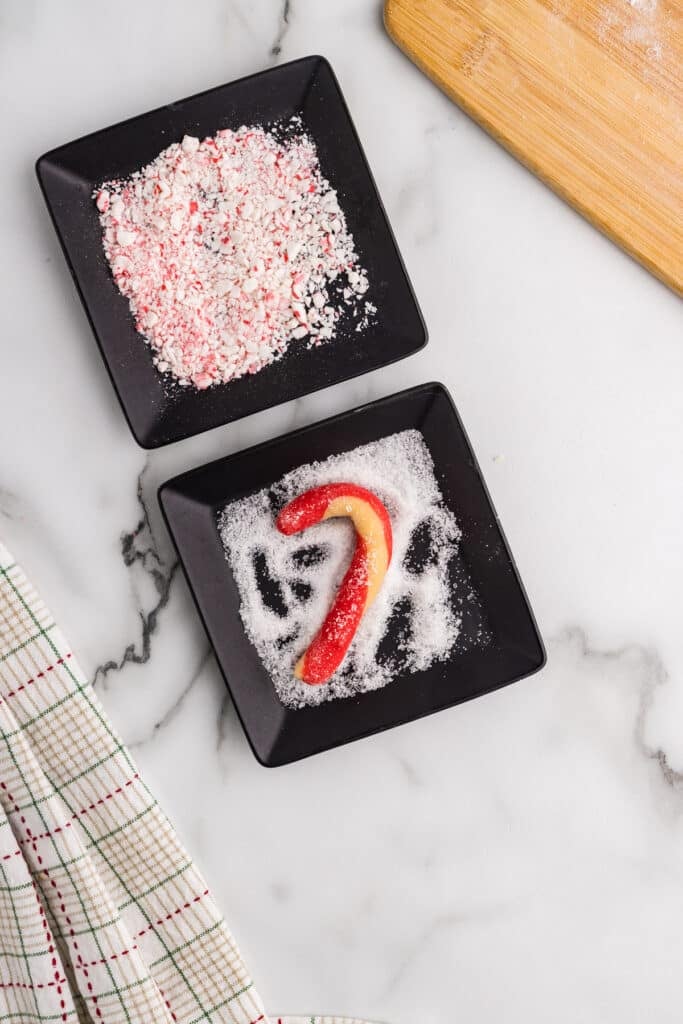 Candy cane shaped resting in a bowl of white sugar with crushed candy pieces.