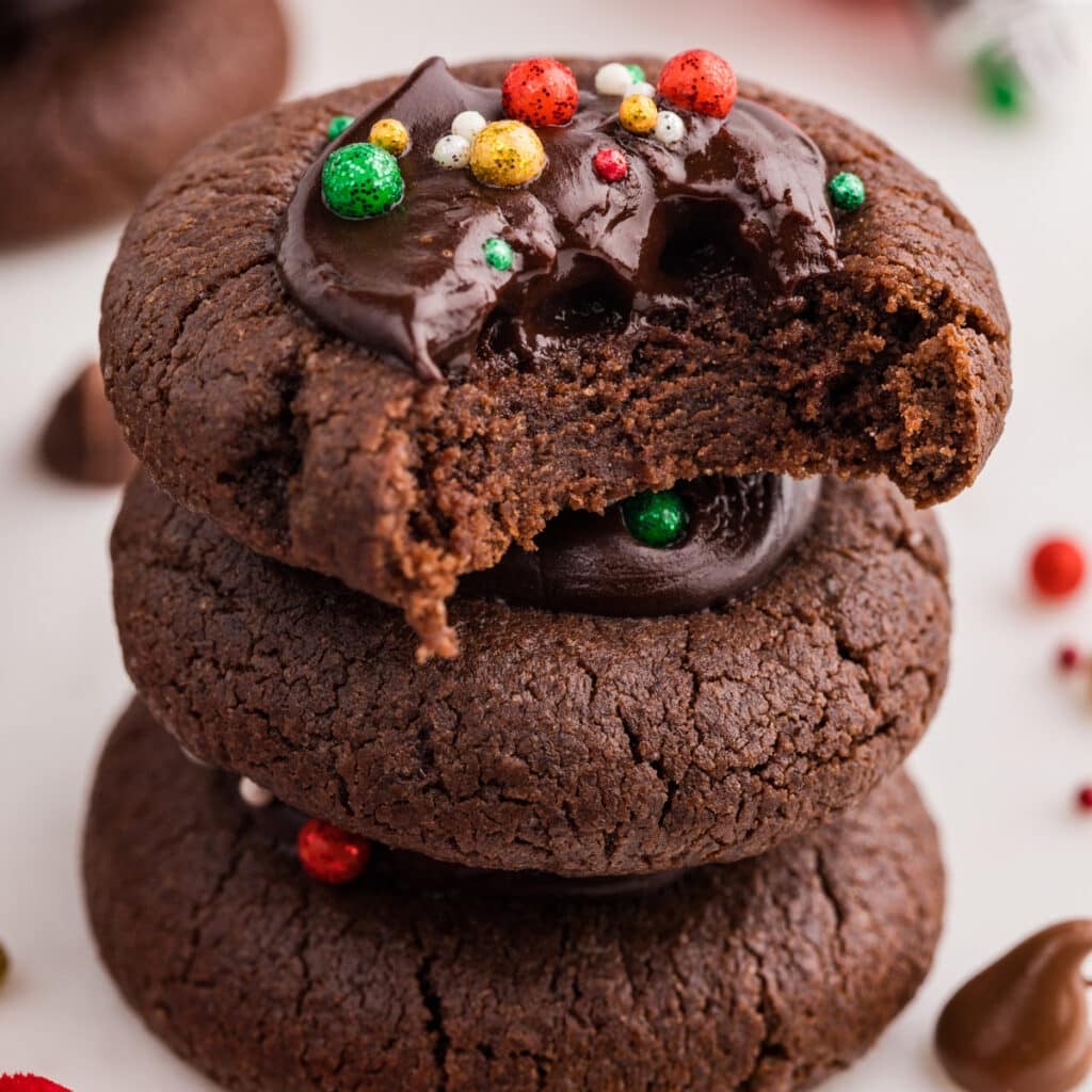 A stack of chocolate thumbprint cookies with chocolate centers and sprinkles, the top one bitten.
