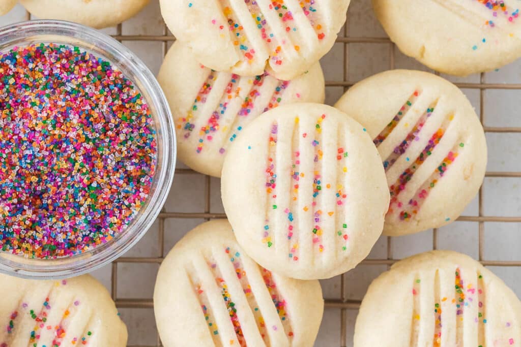 Shortbread cookies cooling on a wire rack with a bowl of rainbow sprinkles.