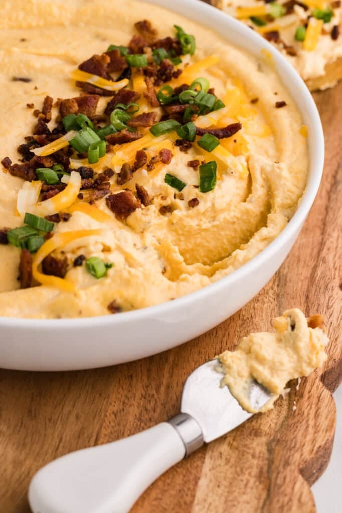 Creamy bacon and cheese dip in a white bowl topped with bacon, shredded cheese, and green onions.