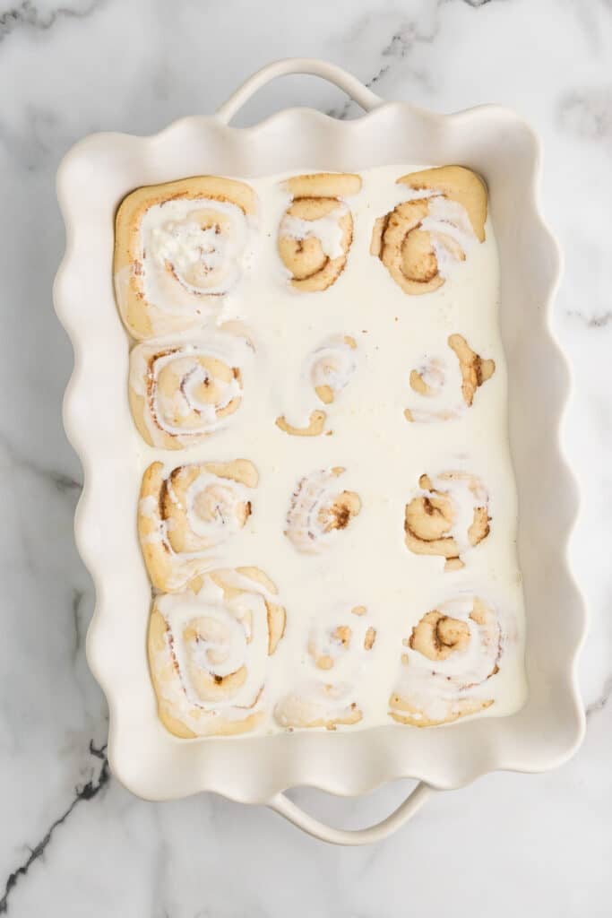Unbaked cinnamon rolls covered in a white liquid in a baking dish.