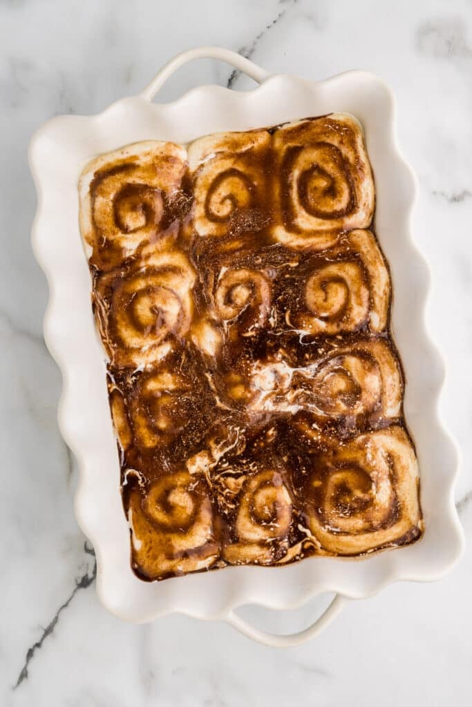 Unbaked cinnamon rolls covered in liquid in a white baking dish.