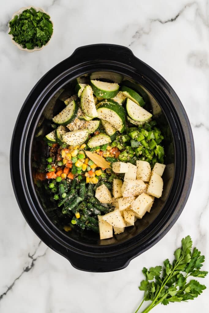 Overhead view of chopped vegetables and seasonings inside a black slow cooker.