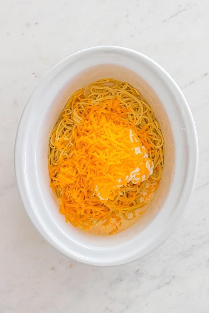 Spaghetti in slow cooker topped with shredded cheddar and dollops of cream cheese.