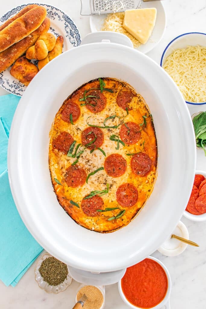 Slow cooker pizza dip inside a white crockpot with pepperoni and basil.