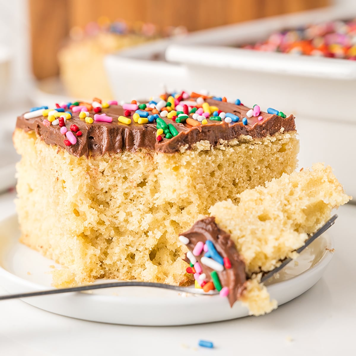 Slice of yellow cake with chocolate frosting and sprinkles on a plate with a fork.